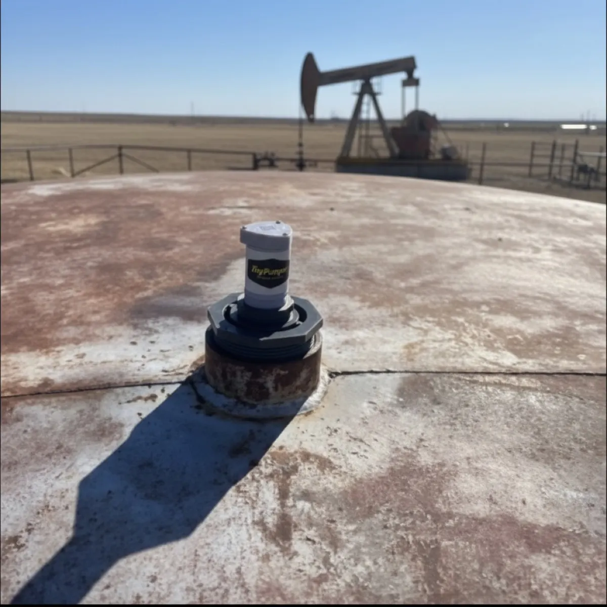 A TinyPumper sensor on a tank hatch in bright daylight, pumpjack on the horizon